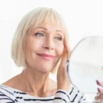 Female older person puts ant aging cream on wrinkles and looks in the mirror