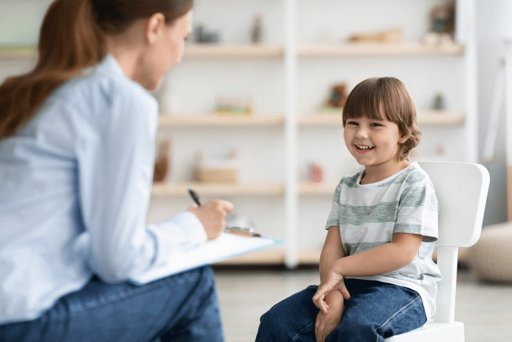 A child smiling while sitting in a chair, engaged in conversation with an adult, who is taking notes.